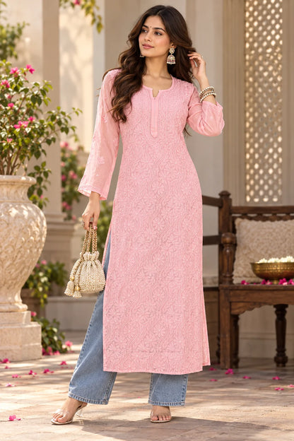 A woman models a light pink long-sleeve kurta with intricate embroidery, paired with light blue wide-leg jeans, holding a small woven handbag in an outdoor courtyard setting.