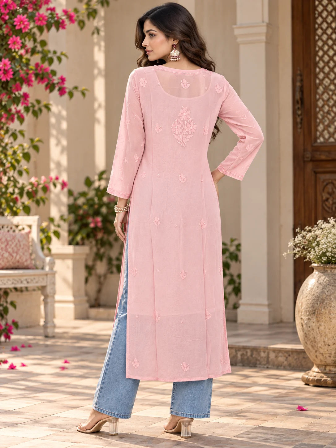 A woman modeling a light peach long kurta with embroidered details, paired with light blue jeans and beige heels, standing in an outdoor courtyard setting.