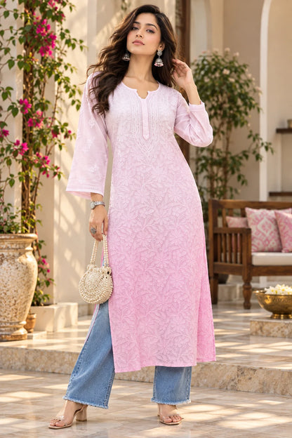 A woman models a light pink embroidered kurta with three-quarter sleeves and a split neckline, paired with blue jeans and holding a small woven handbag in an outdoor courtyard setting.