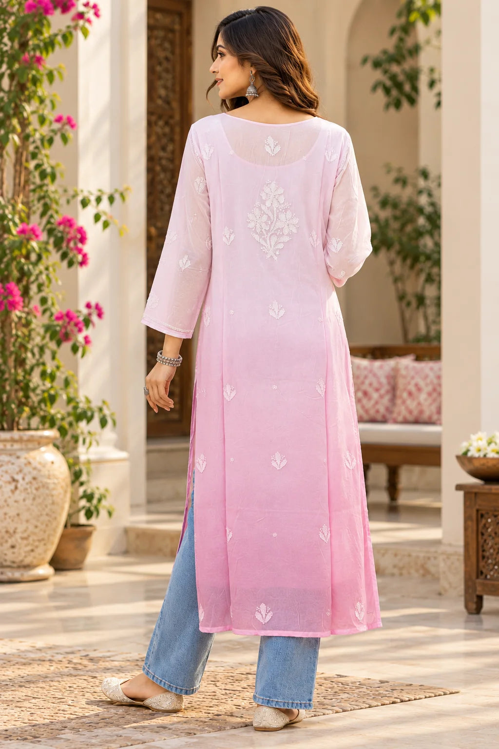 A woman wearing a light pink three-quarter sleeve kurta with white floral embroidery on the back, paired with light blue jeans and white flat shoes, standing in a courtyard setting.