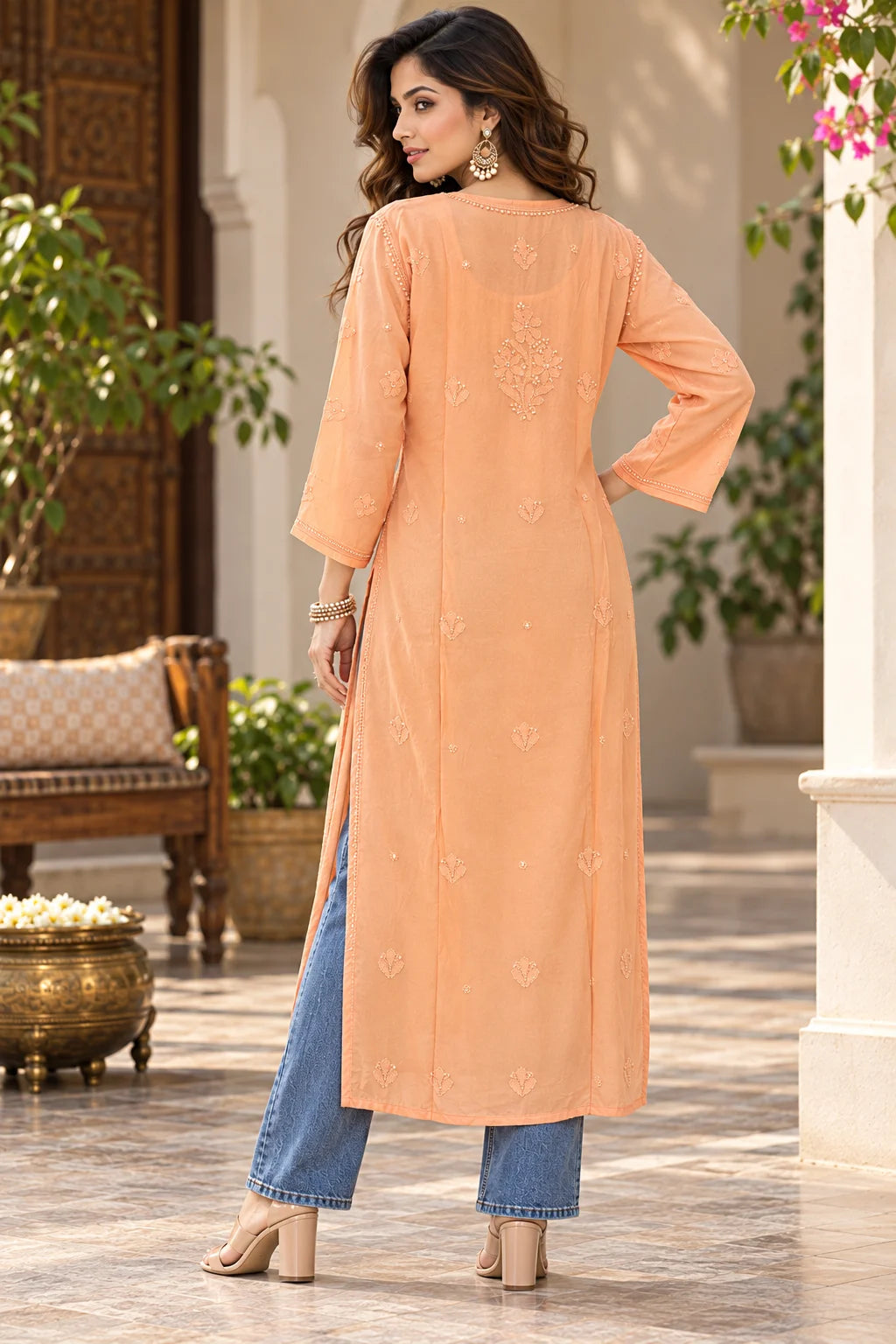 A woman stands in a courtyard wearing a peach-colored embroidered kurta with blue jeans and beige heels, shown from the back.