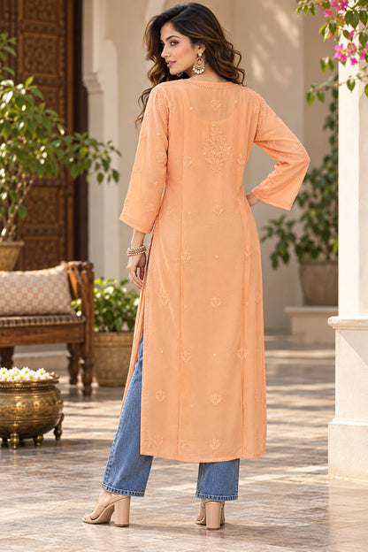 A woman stands in a courtyard wearing a peach-colored embroidered kurta with blue jeans and beige heels, shown from the back.
