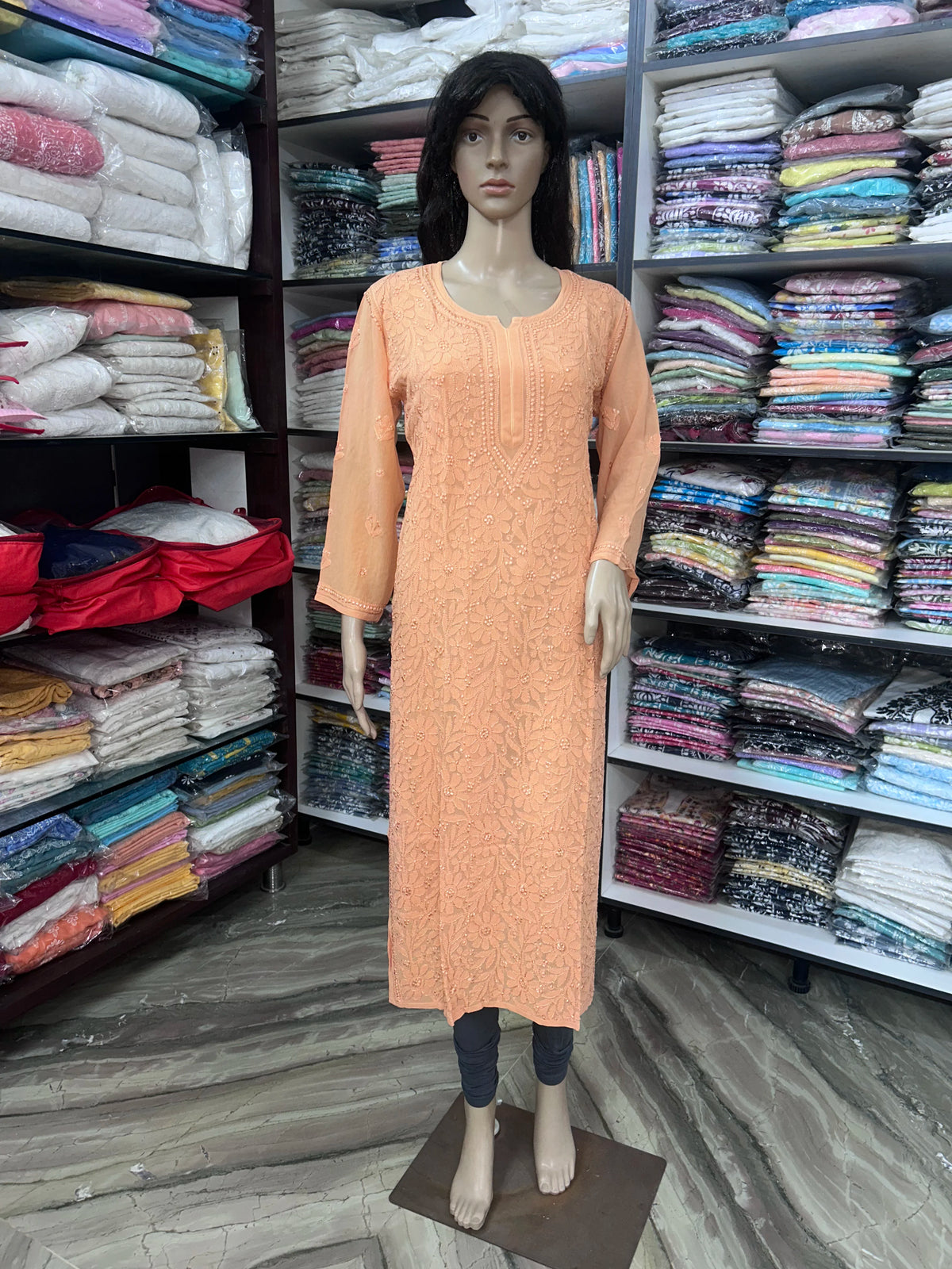 A peach-colored long-sleeve kurta dress displayed on a female mannequin inside a clothing store, with shelves of folded garments visible in the background.