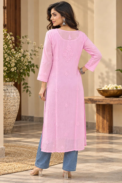 A woman modeling a long pink kurta with subtle embroidery, paired with light blue jeans and beige heels, standing in a warm beige indoor setting.