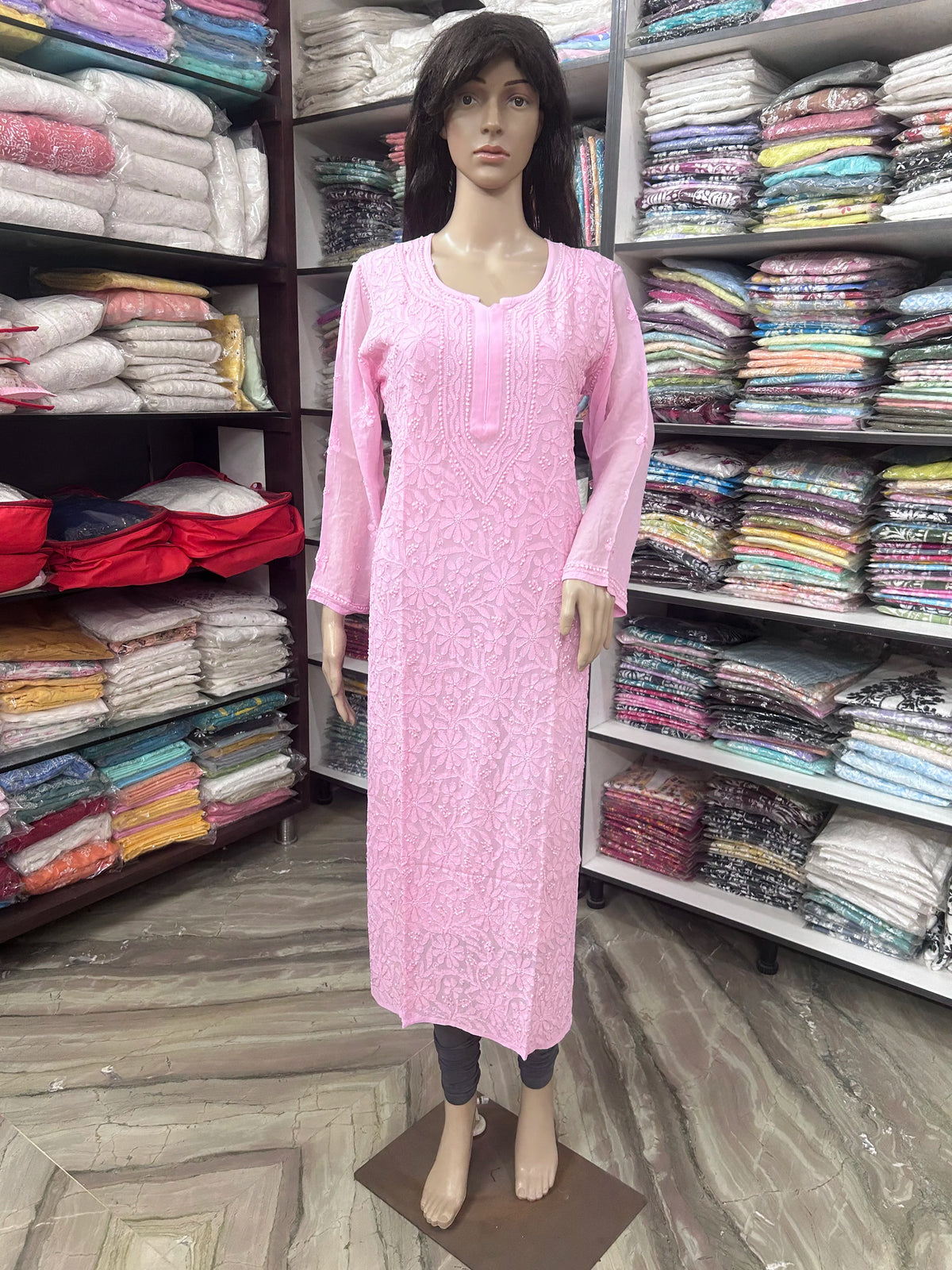 lucknowi chikankari A pink long-sleeve kurta displayed on a female mannequin in a clothing store, with shelves of folded fabrics in the background.