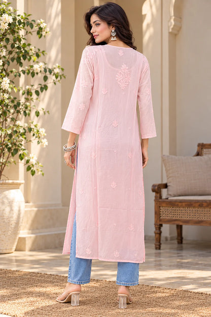 A woman modeling a light pink embroidered kurta with three-quarter sleeves, paired with light blue jeans and beige high-heeled sandals, standing in a sunlit courtyard setting.