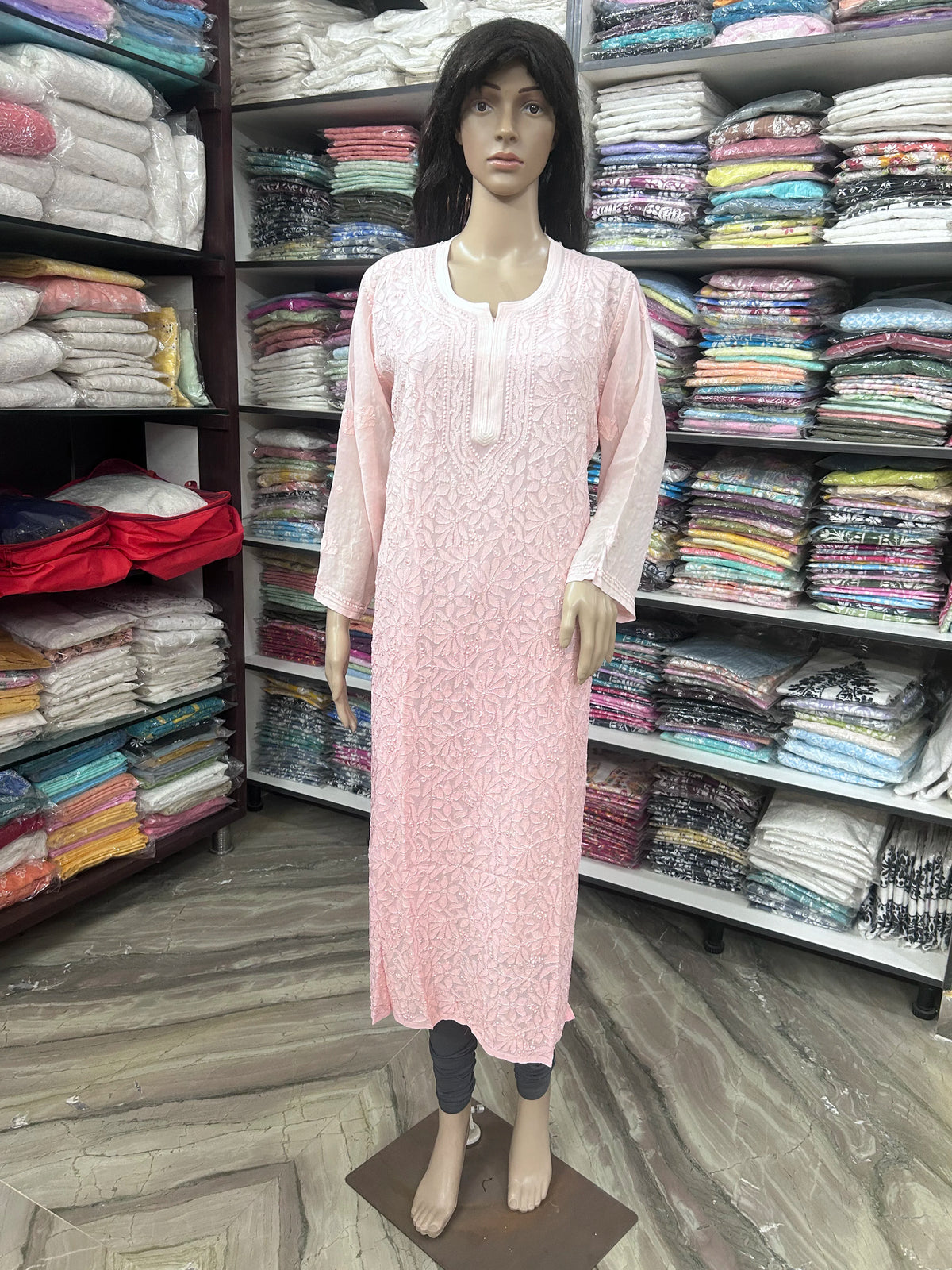 A pink long kurta with a subtle floral jacquard pattern, displayed on a female mannequin in a clothing store. The kurta features a round neckline with a decorative split button placket and three-quarter sleeves.
