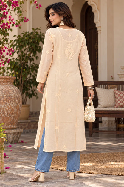 A woman modeling a long peach kurta with subtle embroidery, paired with light blue jeans and beige heels, standing in a warm beige indoor setting.