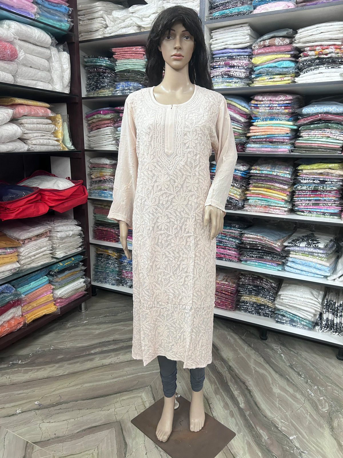 A mannequin wearing a light pink embroidered long kurta with three-quarter sleeves, standing in a clothing store with shelves of folded fabrics in the background.