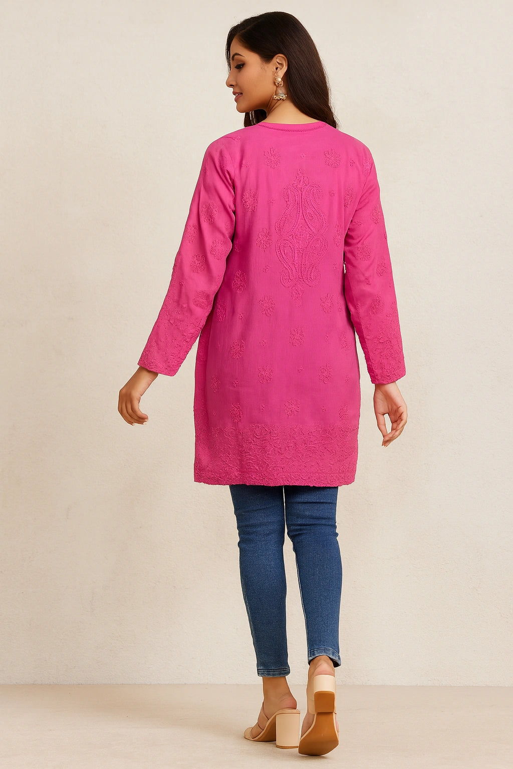 Woman wearing a lucknowi chikankari bright pink long-sleeve top and blue jeans on a beige background