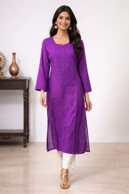 A woman modeling a purple embroidered kurta with white pants and gold sandals, standing in a studio setting with a light-colored wall and a dark wooden side table in the background.
