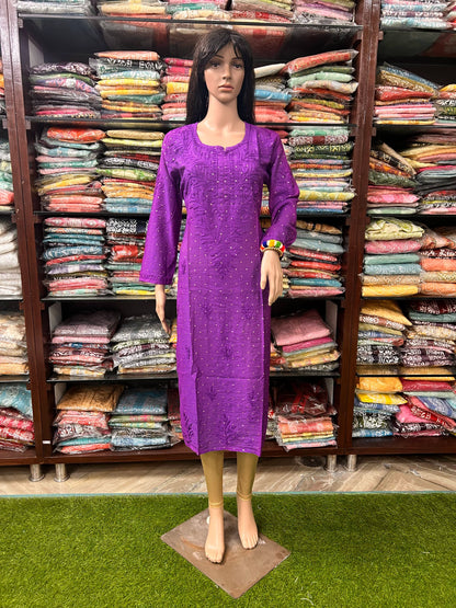 A mannequin wearing a bright purple long-sleeve embroidered dress stands in front of shelves filled with folded colorful garments in a retail store.