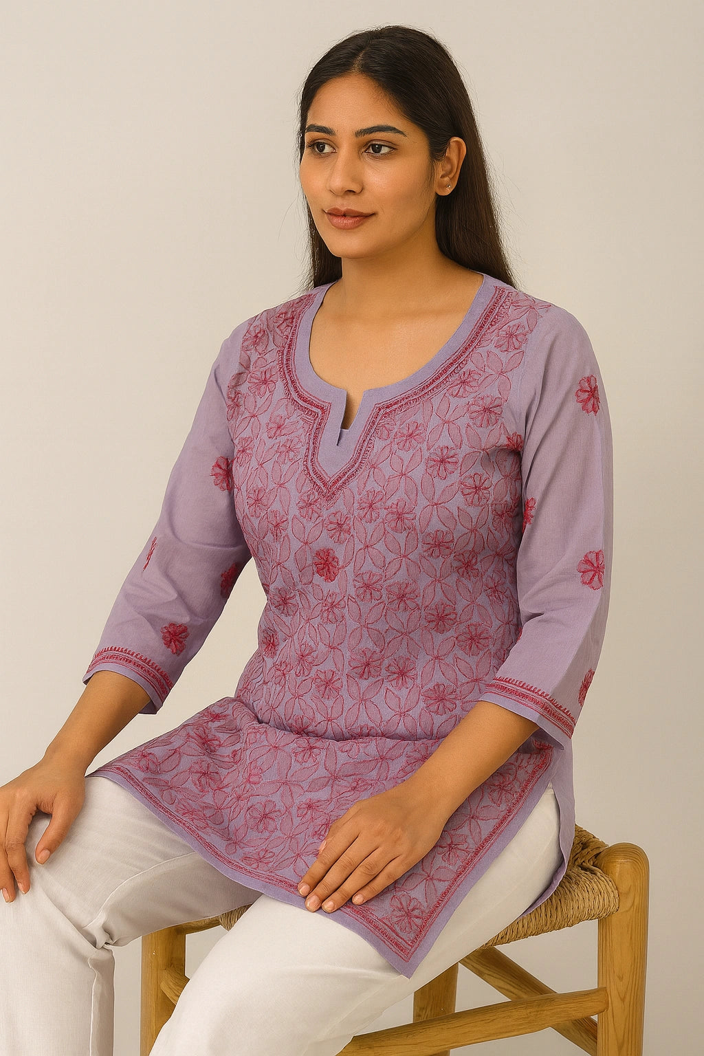 Woman wearing a purple embroidered top sitting on a chair against a plain background