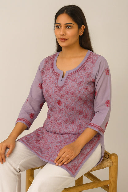 Woman wearing a purple embroidered top sitting on a chair against a plain background