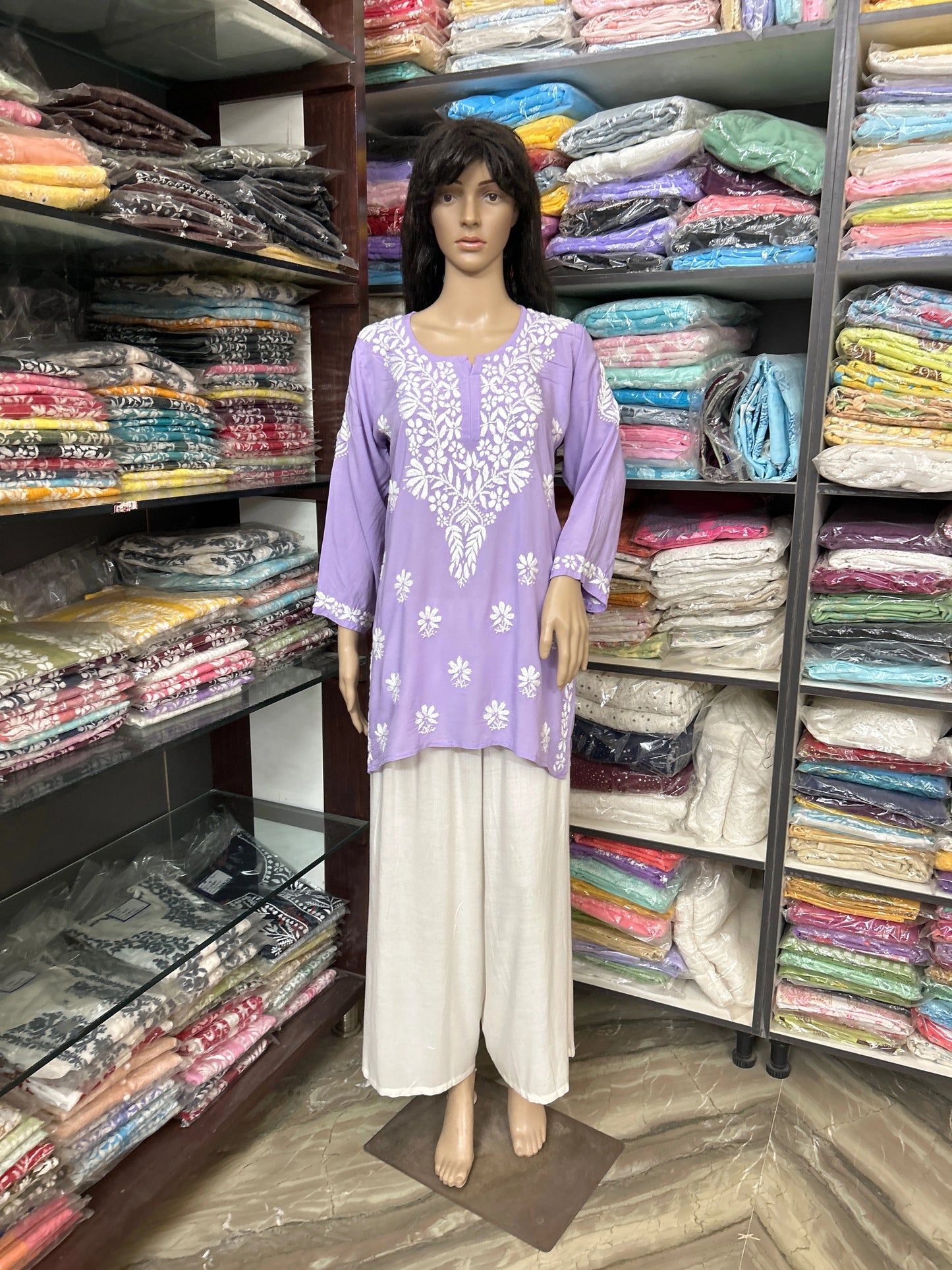 lucknowi chikankari Mannequin wearing a purple top and beige pants in a fabric store with shelves of colorful fabrics.