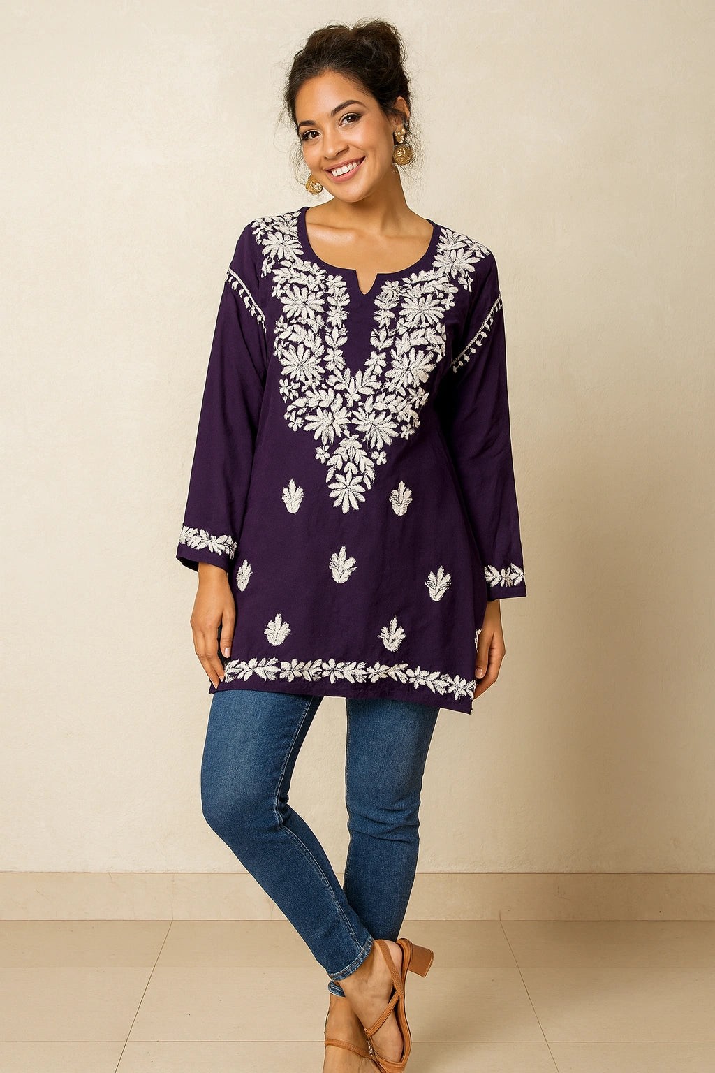 Woman wearing a purple embroidered top and blue jeans against a beige background