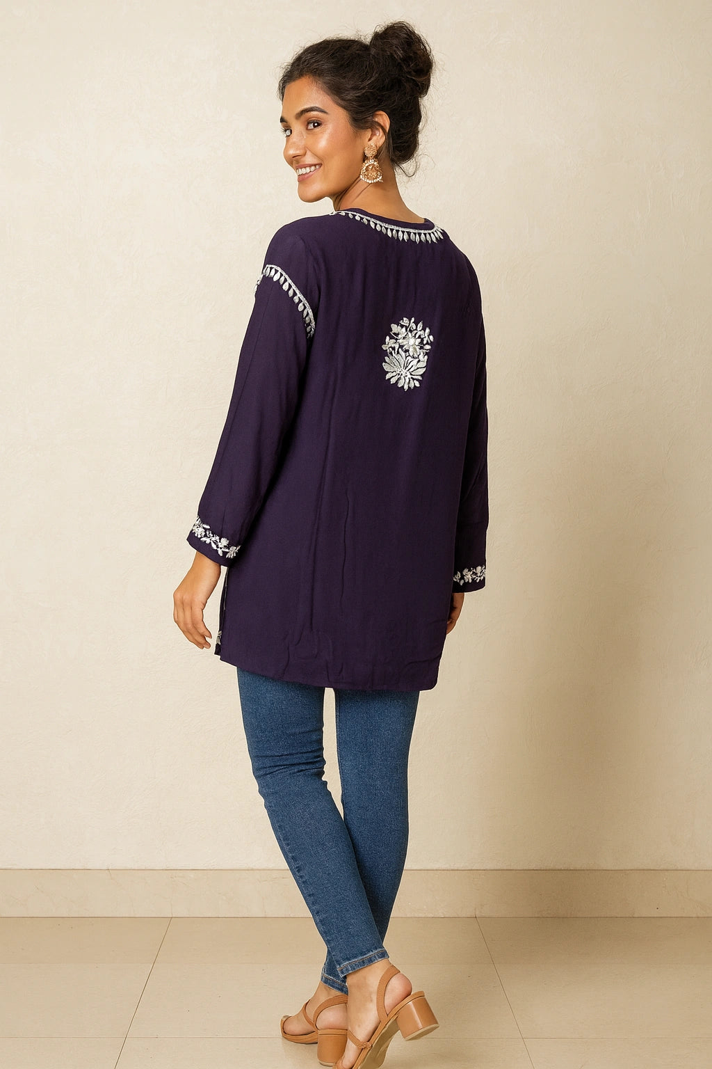 Woman wearing a dark purple long-sleeve top with white embroidery and blue jeans on a beige background