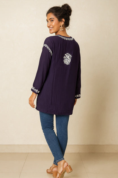 Woman wearing a dark purple long-sleeve top with white embroidery and blue jeans on a beige background