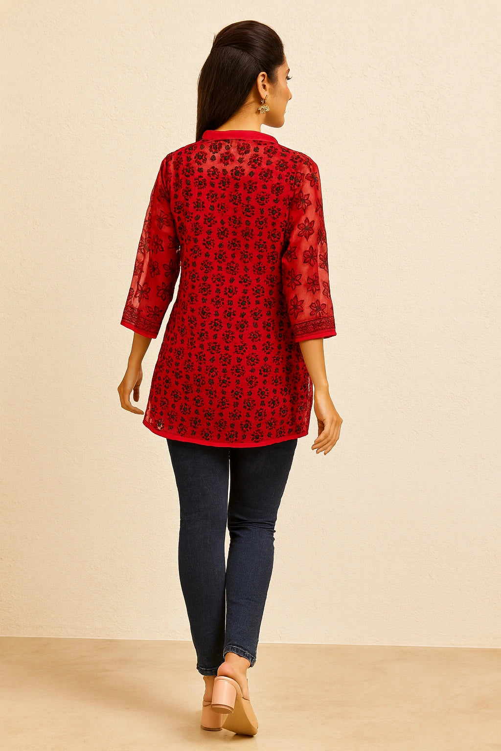 Woman wearing a lucknowi chikankari red floral patterned top and dark blue jeans against a beige background