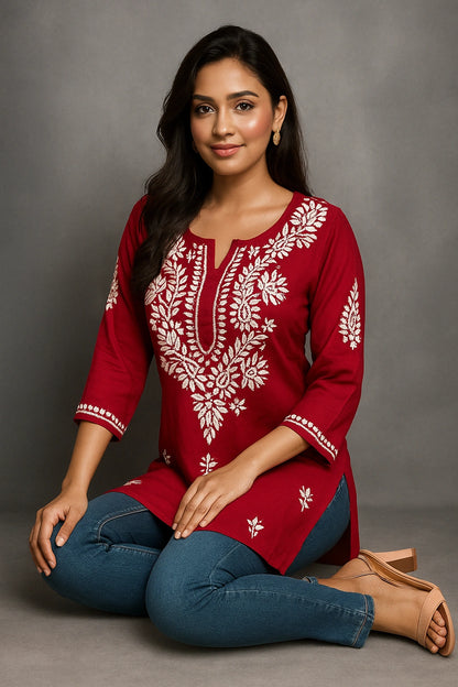 Woman wearing a red embroidered top and blue jeans sitting on the floor against a gray background