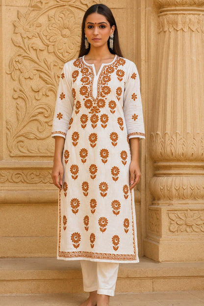 Woman wearing a lucknowi chikankari white kurta with brown floral patterns in front of an ornate beige wall.