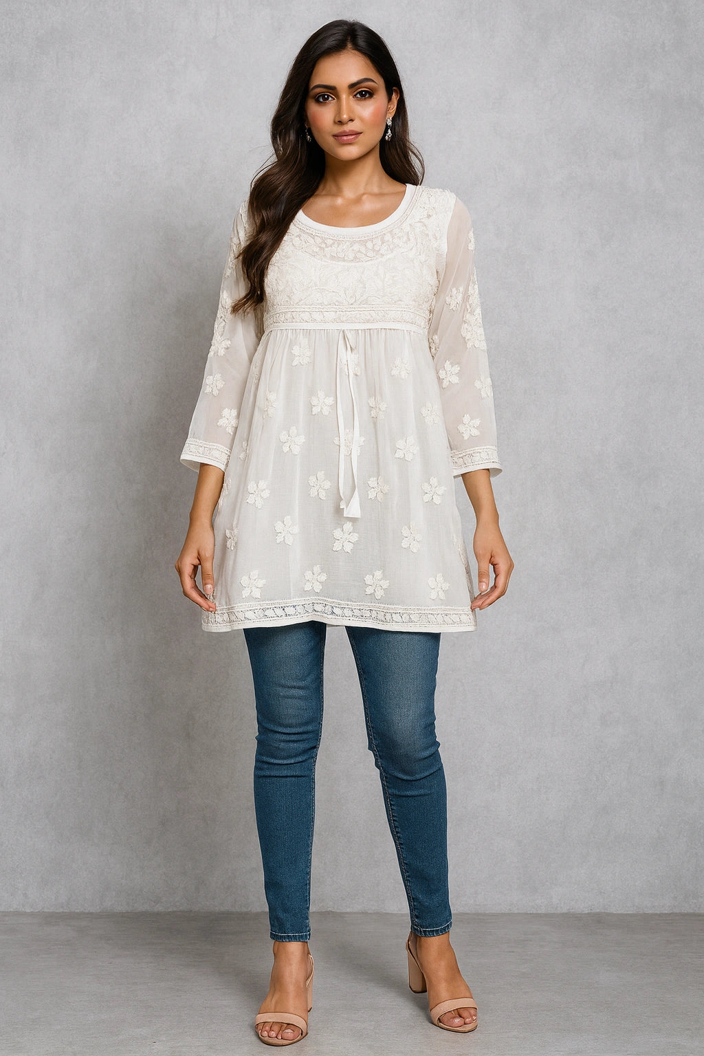 Woman wearing a white embroidered top and blue jeans against a gray background