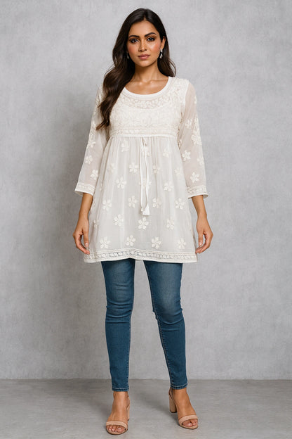 Woman wearing a white embroidered top and blue jeans against a gray background
