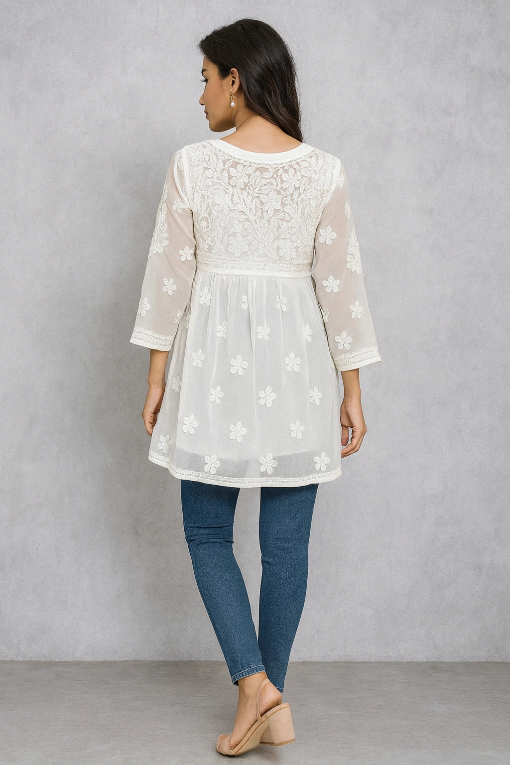Woman wearing a lucknowi chikankari white lace top with sheer sleeves and blue jeans against a gray background