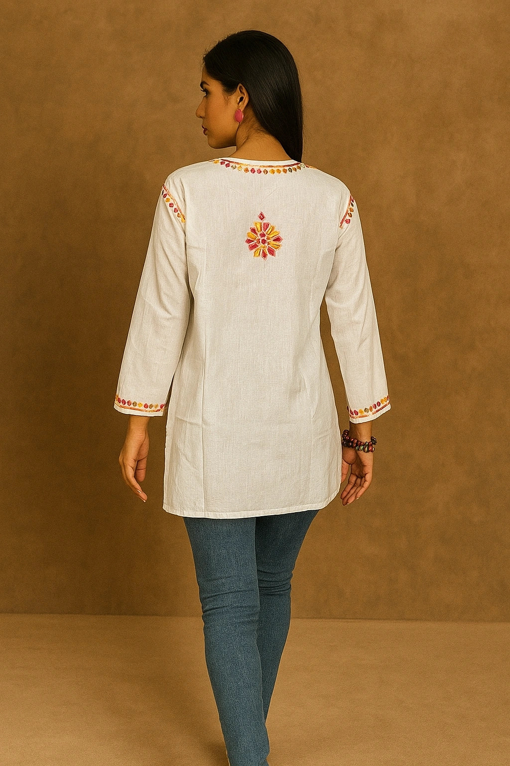 Woman wearing a cream-colored embroidered top with blue pants against a brown background