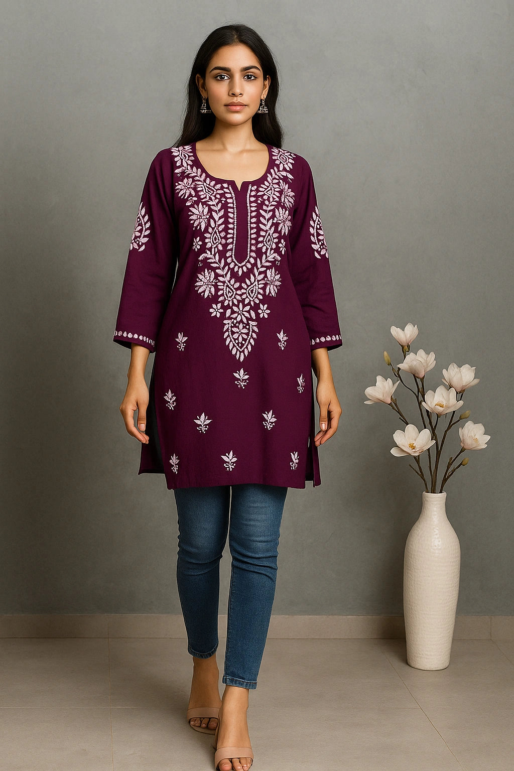 Woman wearing a purple embroidered top with blue jeans standing next to a vase with flowers against a gray background