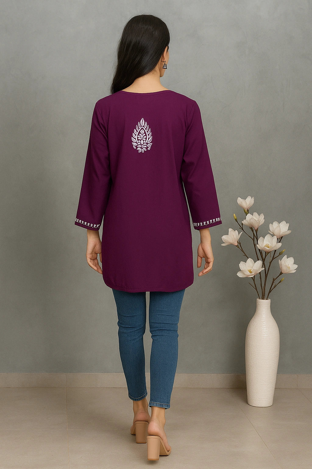 Woman wearing a lucknowi chikankari purple top with a white floral design, standing against a gray wall with a vase of flowers.
