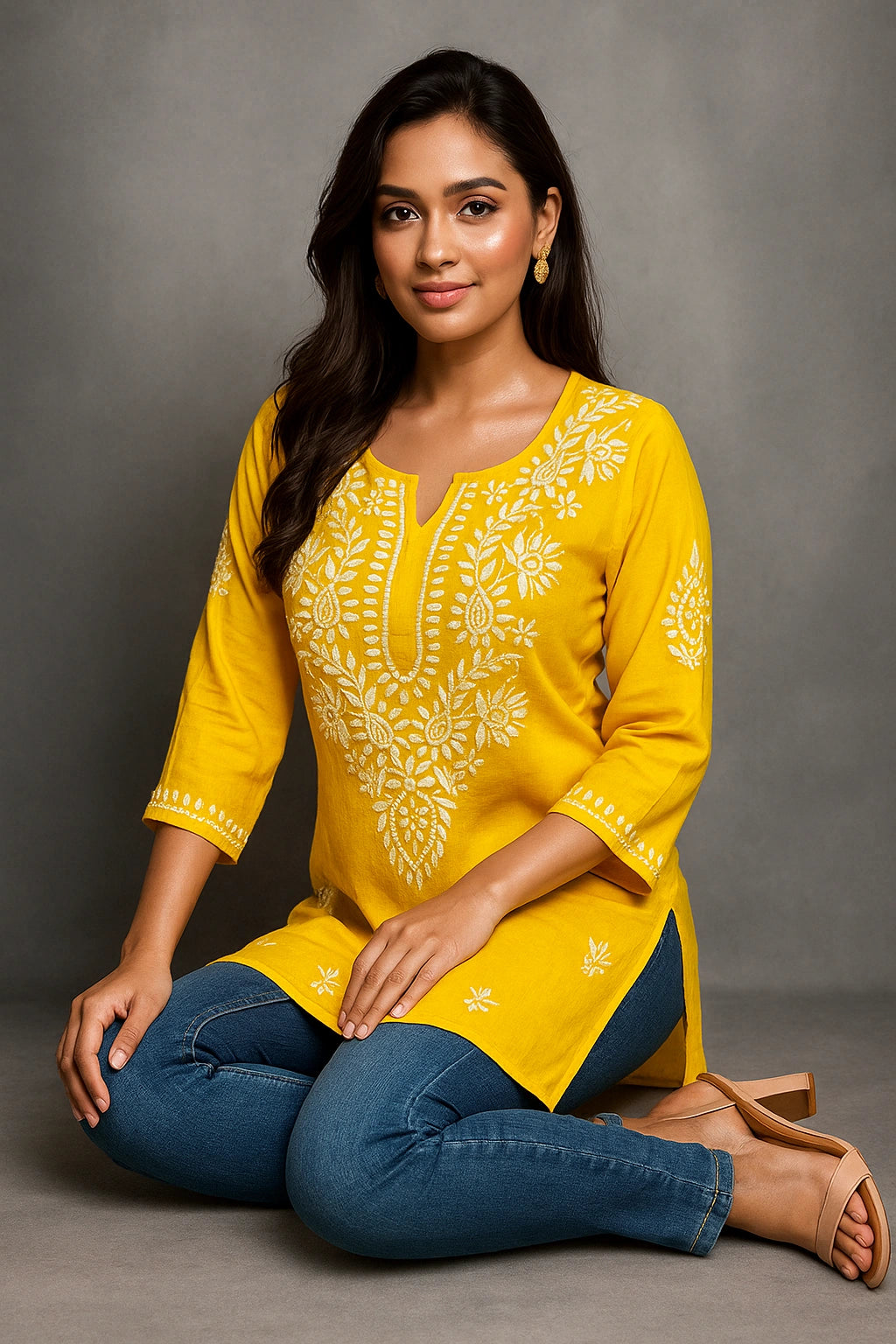 Woman wearing a yellow embroidered top and blue jeans sitting on the floor against a gray background