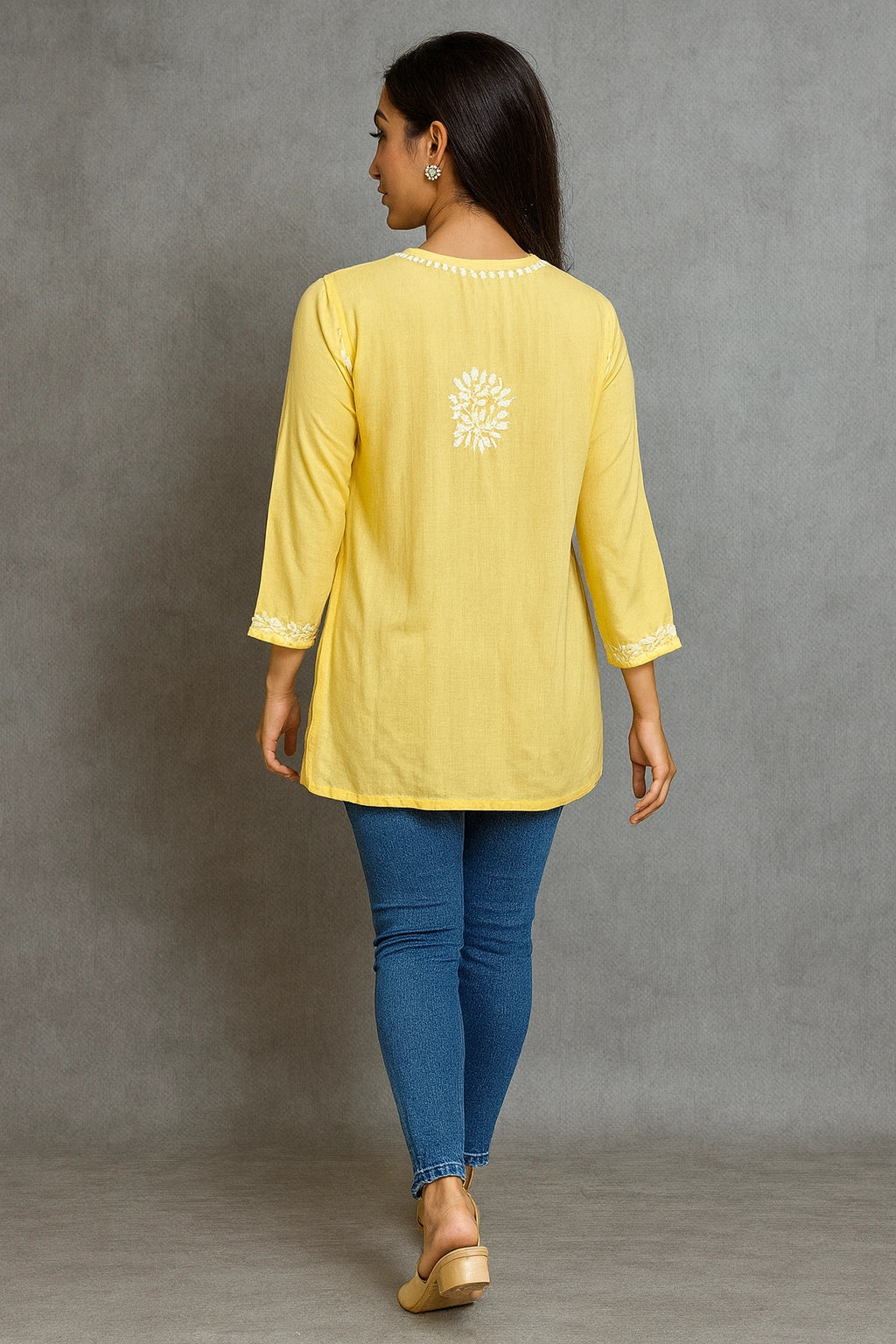 Woman wearing a lucknowi chikankari yellow top with a floral design and blue jeans against a gray background