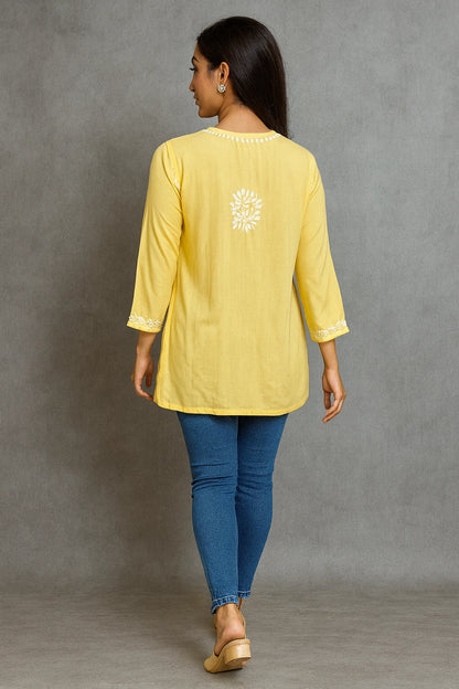 Woman wearing a lucknowi chikankari yellow top with a floral design and blue jeans against a gray background