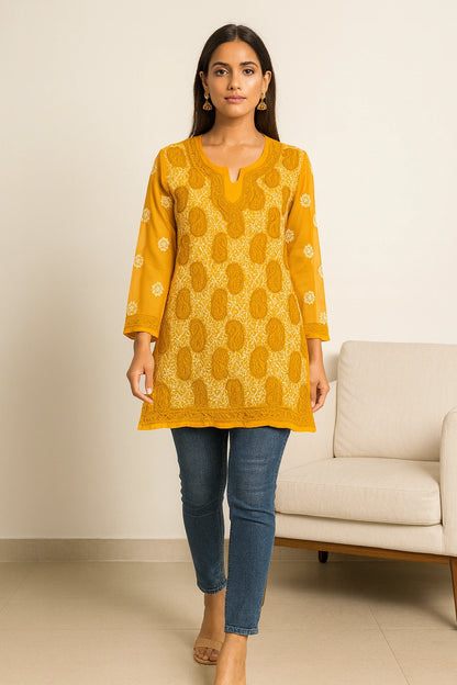Woman wearing a lucknowi chikankari yellow patterned kurta with blue jeans in a minimal indoor setting.