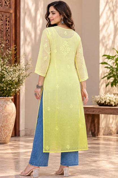 A woman modeling a long yellow kurta with subtle embroidery, paired with light blue jeans and beige heels, standing in a warm beige indoor setting.