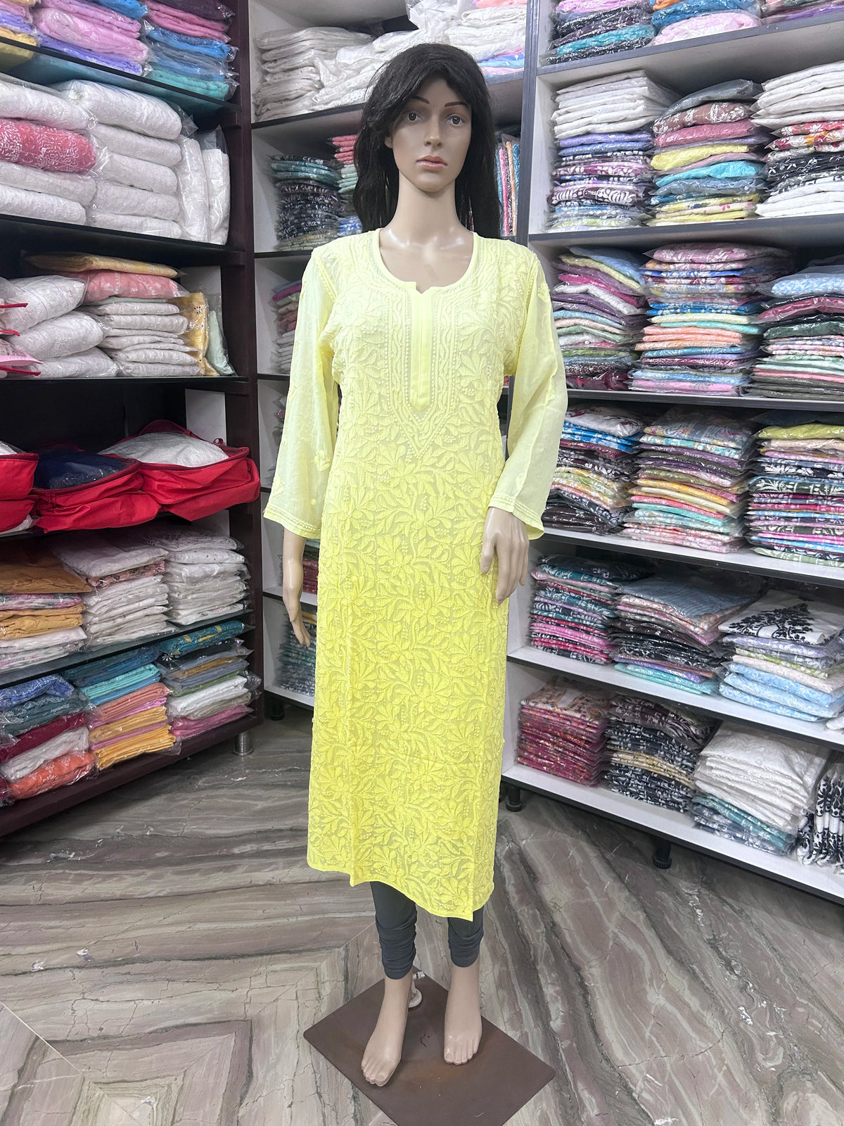 lucknowi chikankari A yellow long-sleeve kurta displayed on a female mannequin in a clothing store, with shelves of folded fabrics in the background.